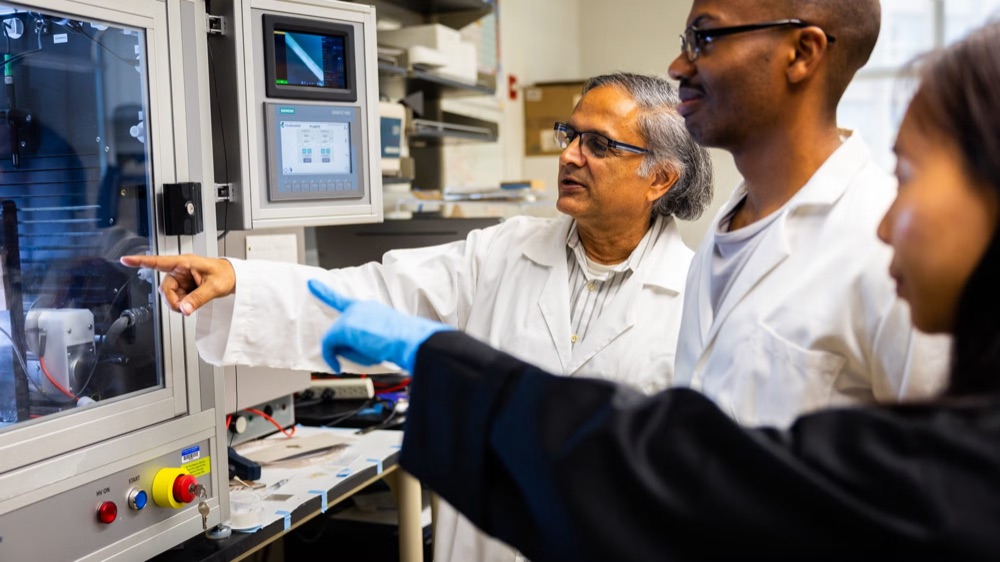 Dr. Makarand (Mak) Paranjape, in a white lab coat, pointing at a piece of
                    laboratory equipment while two researchers in lab coats look on.