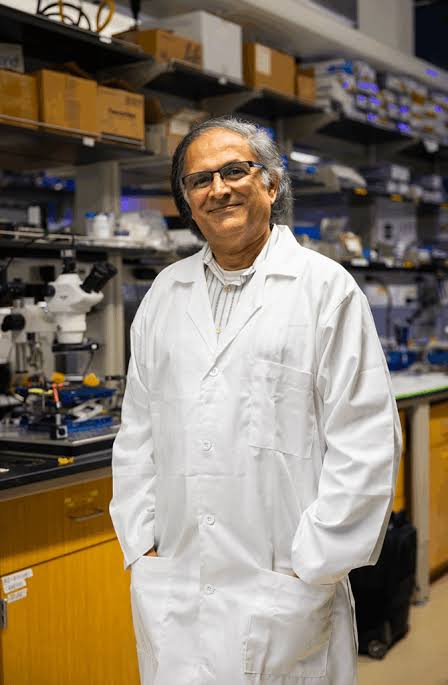 Dr. Makarand (Mak) Paranjape in a white lab coat, standing in the GNuLab clean room beside a microscope and benches of equipment.